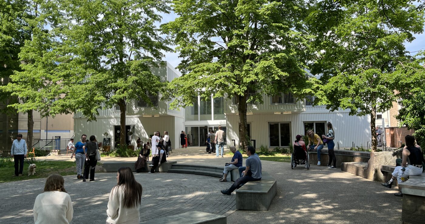 Outdoor courtyard at Hub8 MX in central Cheltenham, with people seated on stone benches beside white modular innovation buildings and trees, the venue for Tuesday's resilience and sovereignty day.