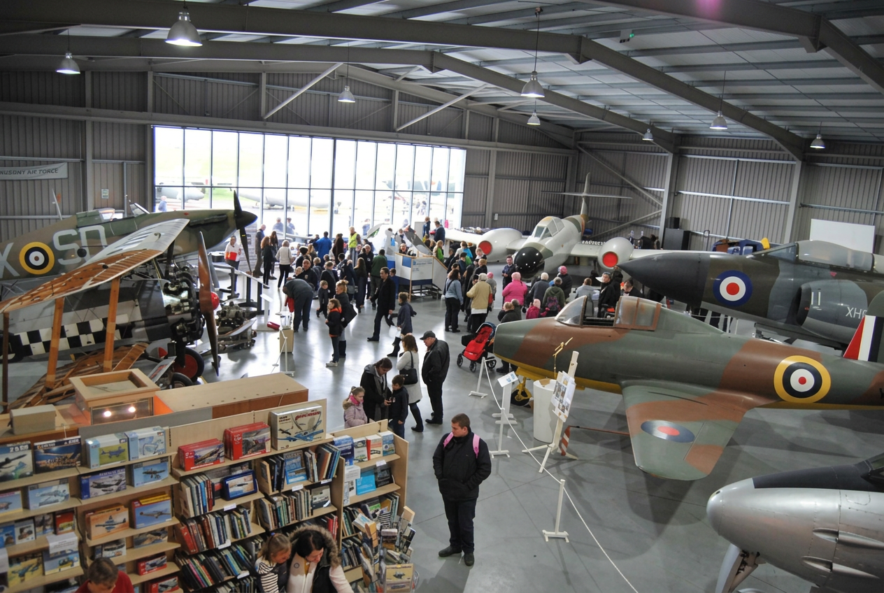 Interior of the Jet Age Museum hangar at Gloucestershire Airport, Staverton, with vintage camouflage and grey jet aircraft on display and visitors walking among the exhibits under a high gabled ceiling, the heritage venue opening Gloucestershire Tech Week on Monday.